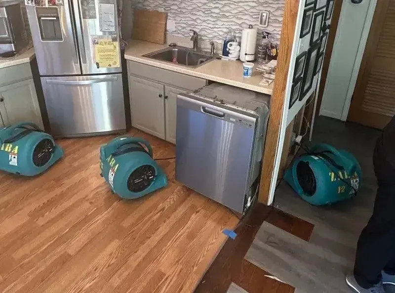 Air movers drying a wood kitchen floor in front of a dishwasher and refrigerator after an appliance leak